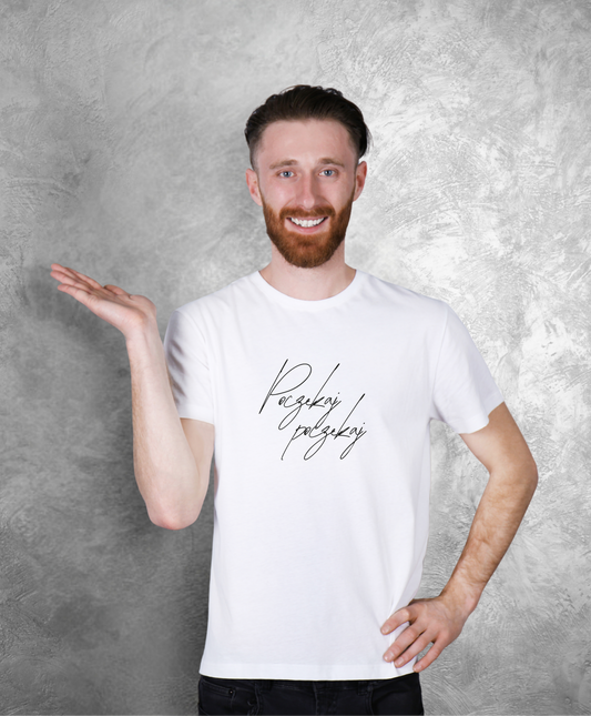 Zdjęcie przedstawia uśmiechniętego mężczyznę stojącego przodem do kamery. Ma na sobie białą koszulkę z czarnym napisem "Poczekaj, poczekaj"