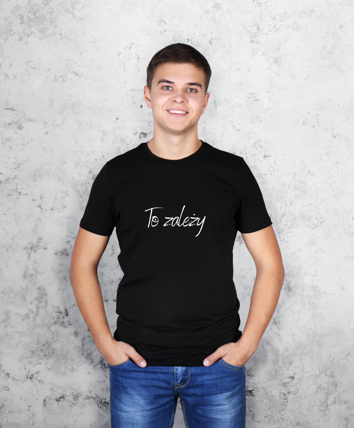 Zdjęcie przedstawia uśmiechniętego mężczyznę, stojącego przodem do kamery, ubranego w czarną koszulkę z białym napisem na przodzie "To zależy"