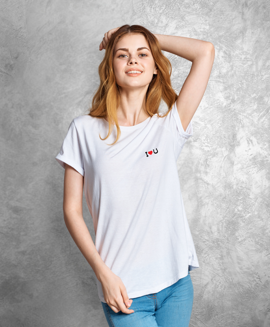 Kobieta stojącą przodem do kamery. Ma na sobie białą koszulkę z czarnym napisem "I love horses" gdzie słowo "love" przedstawione jest jako czerwone serce, a słowo "horses" jako czarna podkowa.  