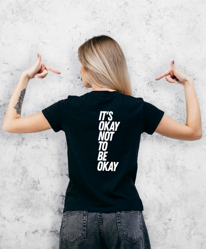 Zdjęcie przedstawia kobietę stojącą tyłem do kamery. Ma na sobie czarną koszulkę z białym napisem "It's ok not to be ok". 