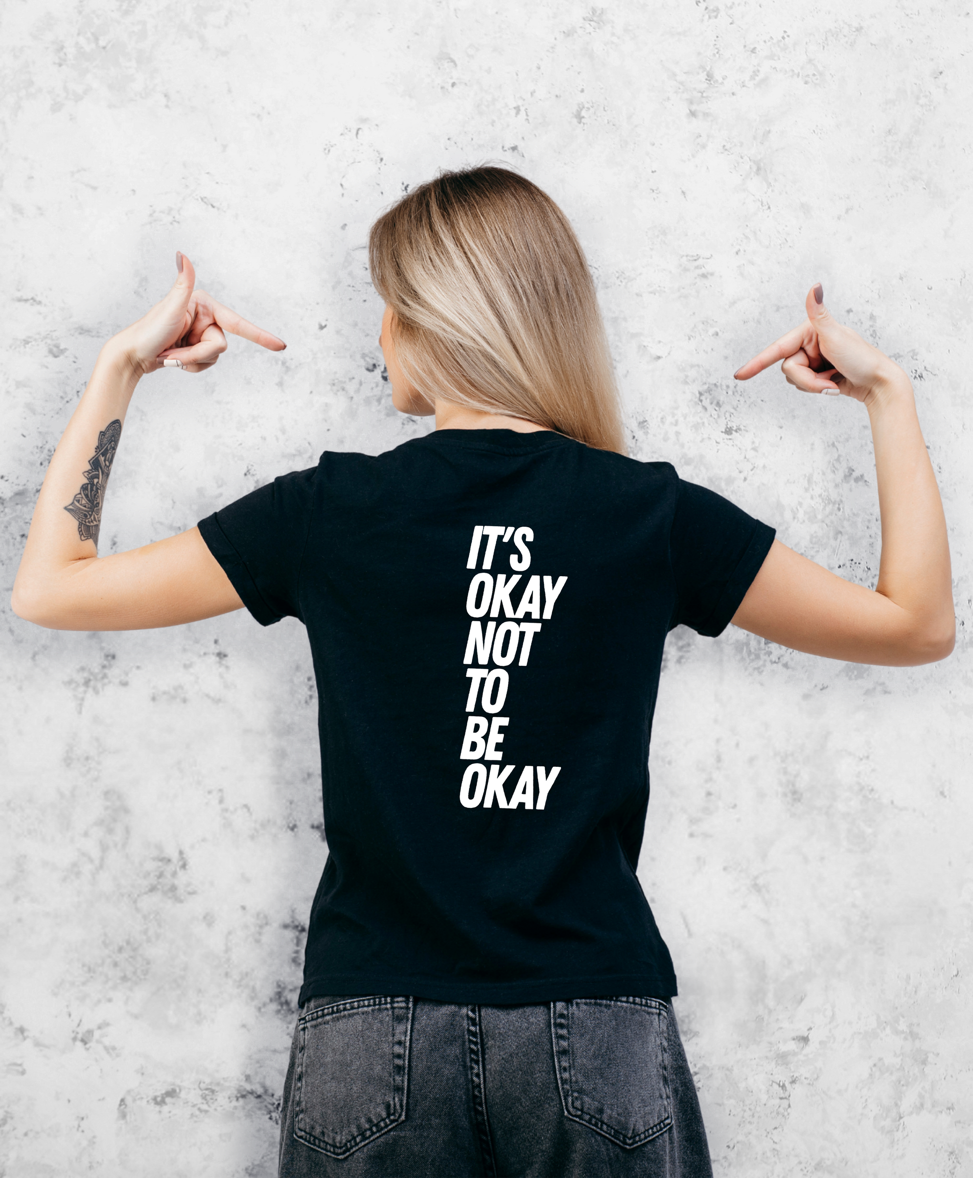 Zdjęcie przedstawia kobietę stojącą tyłem do kamery. Ma na sobie czarną koszulkę z białym napisem "It's ok not to be ok".  