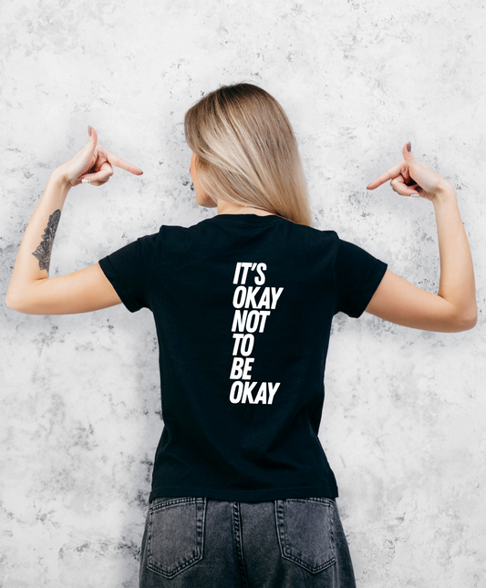 Zdjęcie przedstawia kobietę stojącą tyłem do kamery. Ma na sobie czarną koszulkę z białym napisem "It's ok not to be ok".  