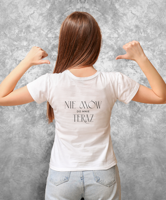 Zdjęcie przedstawia kobietę stojącą tyłem do kamery, ubraną w białą koszulkę z czarnym napisem "Nie mów do mnie teraz" na plecach.