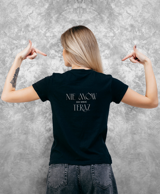 Zdjęcie przedstawia kobietę stojącą tyłem do kamery, ubraną w czarną koszulkę z białym napisem "Nie mów do mnie teraz" na plecach.
