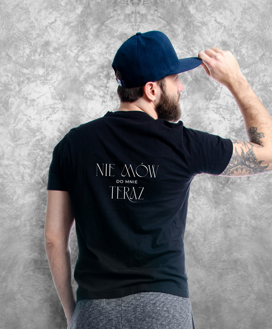 Zdjęcie przedstawia mężczyznę stojącego tyłem do kamery, ubranego w czarną koszulkę z białym napisem "Nie mów do mnie teraz" na plecach.