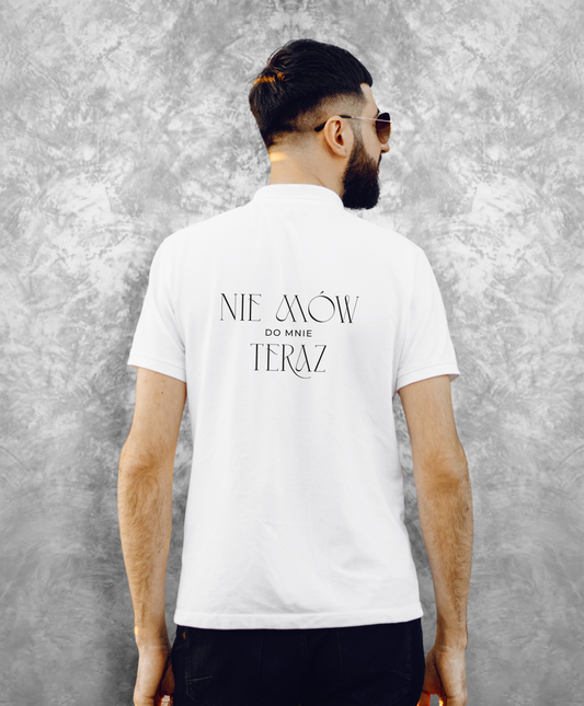 Zdjęcie przedstawia mężczyznę stojącego tyłem do kamery, ubranego w białą koszulkę z czarnym napisem "Nie mów do mnie teraz" na plecach.