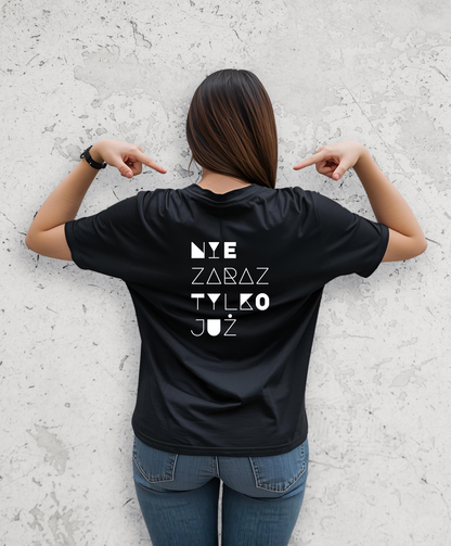 Kobieta stojąca tyłem do kamery. Ubrana w czarny t-shirt z białym napisem na plecach "Nie zaraz, tylko juz"