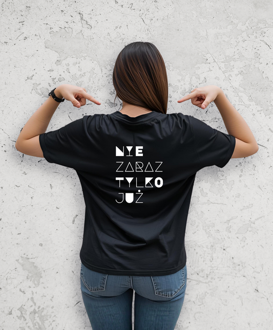 Kobieta stojąca tyłem do kamery. Ubrana w czarny t-shirt z białym napisem na plecach "Nie zaraz, tylko juz"