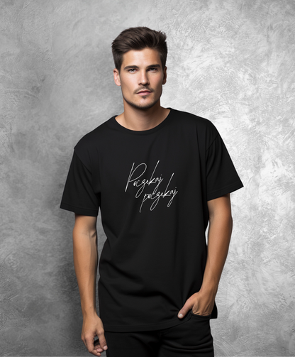 Zdjęcie przedstawia mężczyznę stojącego przodem do kamery. Ma na sobie czarną koszulkę z białym napisem "Poczekaj, poczekaj"  