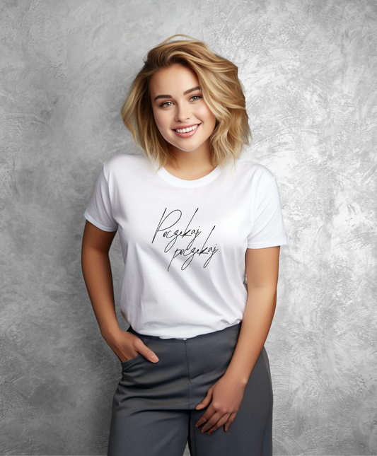 Zdjęcie przedstawia uśmiechniętą kobietę stojącą przodem do kamery. Ma na sobie białą koszulkę z czarnym napisem "Poczekaj, poczekaj"  