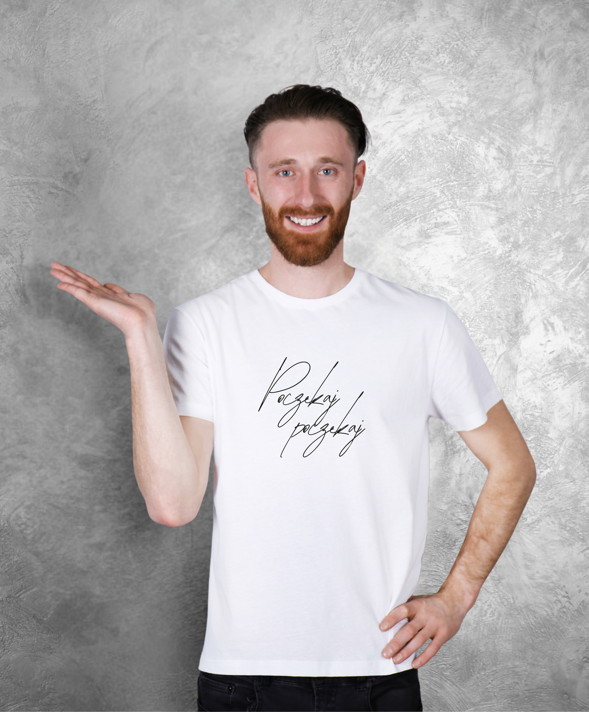 Zdjęcie przedstawia uśmiechniętego mężczyznę stojącego przodem do kamery. Ma na sobie białą koszulkę z czarnym napisem "Poczekaj, poczekaj"  