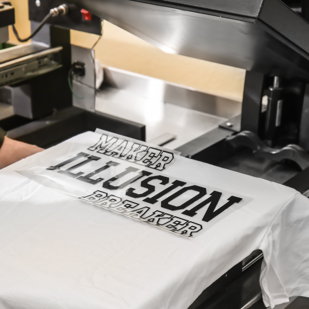 zdjęcie przedstawia białą koszulkę, na którą nałożona jest naklejka z tekstem. Koszulka leży na maszynie, która przykleja napis