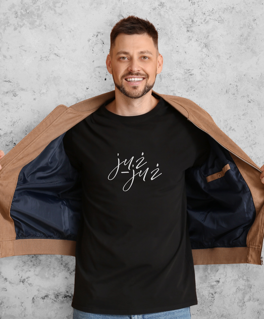 Uśmiechnięty mężczyzna stojący przodem do kamery, ubrany w czarną koszulkę z białym napisem "już już"