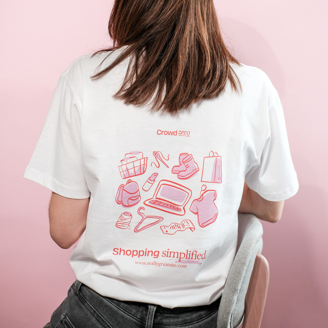 zdjęcie przedstawia kobietę na różowym tle, siedzącą na krześle tyłem do nas, ma na sobie białą koszulkę z różowymi grafikami przedstawiającymi m.in. komputer, wieszak, laptop, koszulkę, plecak.